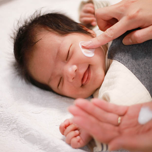 Einem Baby mit vollem dunklen Haar wird das Gesicht eingecremt.