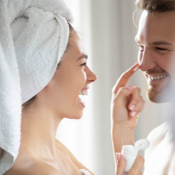 Profilaufnahme einer Frau ihren Haaren in ein Handtuch gewickelt, die die Nase eines Mannes mit etwas Creme auf ihrem Finger anstuppst.