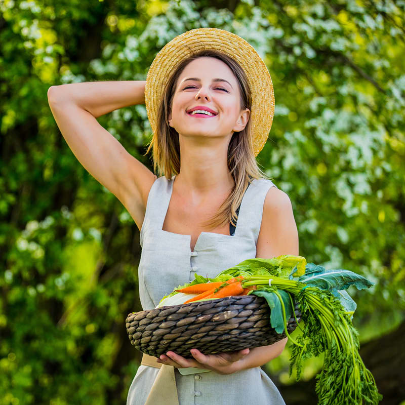 Eine Frau mit Strohhut hält einen Gemüsekorb vor sich und lächelt.