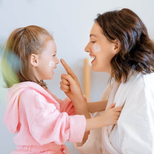 Profilaufnahme einer Mutter und ihrer Tochter; beide haben einen Klecks Creme auf ihrer Wange und lächeln sich an.